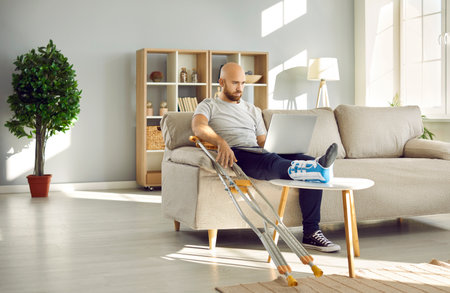 Sick businessman with broken leg in bandage sit on sofa at home work on laptop online. Unhealthy man with foot injury in splint use computer during leave. Trauma and rehabilitation. Medicine concept.の写真素材