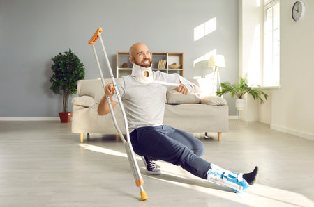 Happy man with multiple physical injuries having fun while recovering at home. Crazy, funny male patient with broken neck, sprained foot and injured arm moving and dancing with crutch in living roomの写真素材