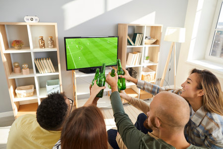 Young friends clinking beer bottles while watching football on TV. Happy multiethnic male and female fans sitting in the living room, watching soccer, drinking beer, and having a good time togetherの写真素材