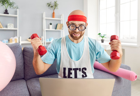 Funny man in retro sportswear holding dumbbells and looking at laptop. Bearded guy sitting on sofa doing aerobics. Concept home training, live streaming, sports person, funny picturesの写真素材