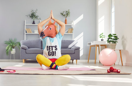 Extraordinary approach to yoga. Man in horse mask is doing funny yoga and performing asana in cozy home interior. Eccentric man sits in lotus position on yoga mat with horse mask on his head.の写真素材