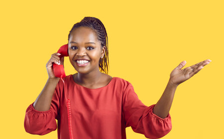 Joyful young African American woman hears good news while talking on landline retro telephone on yellow background. Smiling woman with red headset holding her hand emphasizing copy space.の写真素材