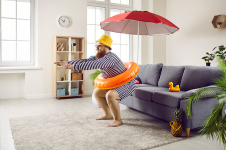 Funny guy staying at home and having cool fake beach holiday in living room. Happy man in hat and striped swimsuit pretends to jump in imaginary ocean waters. Summer vacation conceptの写真素材
