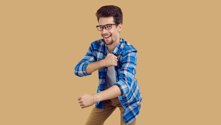 Happy student in casual clothes dancing at a student party. Portrait of a cheerful, joyful young man in a blue plaid shirt and glasses dancing isolated on a beige banner backgroundの写真素材