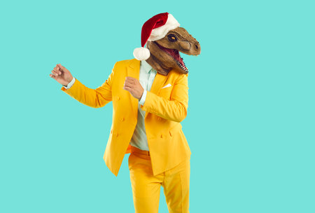Cool freaky man with dinosaur head wearing santa hat having fun, happy and dancing at christmas party. Man in yellow suit and with animal head isolated on light blue background. Christmas banner.の写真素材