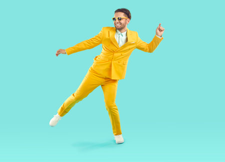 Excited confident stylish young man dancing with happy smile. Full length photo of positive man in sunglasses and trendy yellow suit having fun standing on one leg over light blue backgroundの写真素材