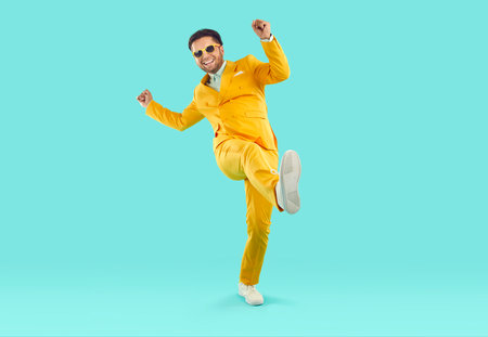 Full body shot of a happy, cheerful man wearing a trendy, stylish yellow suit and sunglasses having fun and dancing on a turquoise blue studio background. Party and fashion conceptの写真素材