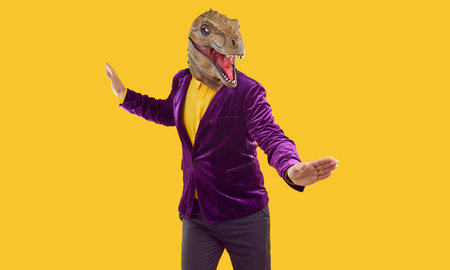 Eccentric funny showman in rubber dinosaur mask, yellow shirt and velvet jacket dancing with wide hands isolated on yellow background. Party, masquerade with costumes. Funky man using animal style.の写真素材