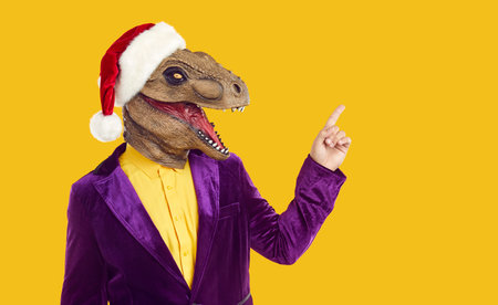 Funny excited dinosaur guy in Christmas cap pointing finger to side on yellow studio background. Man with bizarre dino head advertising holiday or inviting to festive fancy dress masquerade partyの写真素材