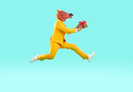Eccentric man in horse mask jumping with gift box. Side view shot of man wearing yellow suit and animal head mask jumping in air carrying present box with red bow on light blue backgroundの写真素材