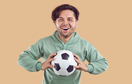 Soccer fan happy excited crazy smiling wondered young man in green sweatshirt holding football ball in hands isolated on beige background and looking at camera. Victory, goal, game, fan concept.の写真素材