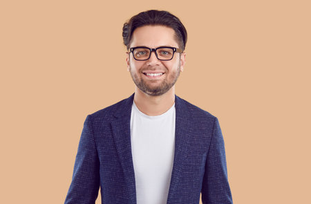 Portrait of young confident unshaven brunet smiling man in blue jacket, white t-shirt and glasses isolated on beige background. Businessman, entrepreneur in casual suit is looking at camera.の写真素材