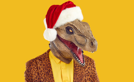 Portrait of man in funny unusual Christmas costume. Headshot of strange guy wearing bizarre dinosaur monster mask, red Santa Claus hat and funky leopard jacket isolated on yellow color backgroundの写真素材