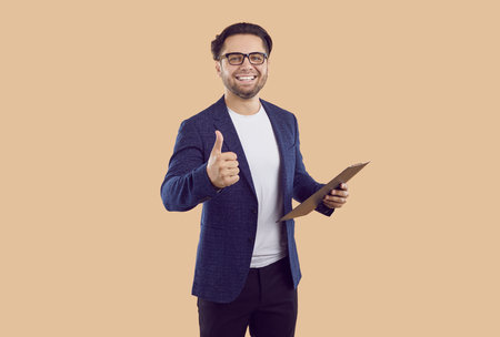 Love my job. Studio shot of successful business agent or sales manager. Happy confident man in suit and glasses standing isolated on beige background, holding clipboard, showing thumbs up and smilingの写真素材