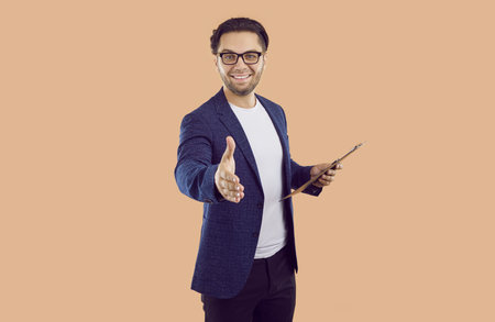 You are hired. Friendly male employer wants to shake your hand congratulating you on getting new job. Smiling businessman in suit holding clipboard reaching out to camera isolated on beige background.の写真素材