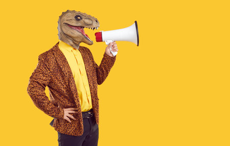 Creative man with dinosaur head makes loud advertisement with help of loudspeaker. Eccentric man in dinosaur mask and leopard print jacket is loudly announcing discounts on orange background.の写真素材