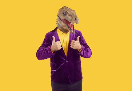 Funny showman in rubber dinosaur mask and purple velvet jacket and yellow shirt showing thumbs up on yellow background. Banner for advertisement, marketing with funky man. Party, masquerade concept.の写真素材