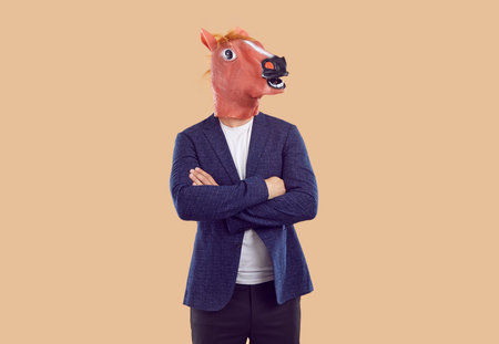 Funny man in rubber horse mask, casual clothes blue blazer, pants and white t-shirt, standing with crossed on chest arms on beige background. Body language, fun, party, masquerade, costume concept.の写真素材