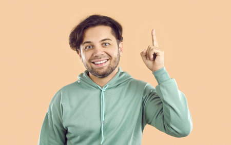 Portrait of attractive smiling happy young man in casual clothes looking at the camera and pointing up with his finger isolated on studio beige background. People emotions concept.の写真素材