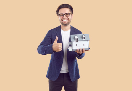 Man real estate agent in glasses, casual blue jacket, white t-shirt and jeans with wooden house in hand smiling showing thumb up on beige background. Sale of real estate, advertisement banner concept.の写真素材