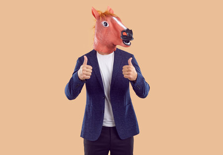 Creative studio shot of man wearing business suit and funny animal masquerade horse mask with silly face expression standing isolated on beige background and giving thumbs upの写真素材