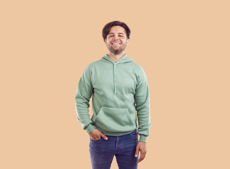 Studio portrait of happy man in casual clothes. Young man in mint green hoodie and blue jeans standing with his hand in pocket isolated on beige background. Casual fashion conceptの写真素材