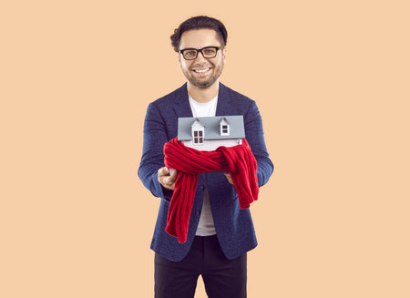 Studio portrait of a happy male real estate agent with a little house. Smiling man in a suit and glasses isolated on a beige background and holding a house wrapped in red cloth. Property sale conceptの写真素材