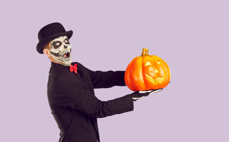 Crazy amazed man in halloween black costume and hat with pumpkin in hands and scary skeleton make-up on purple background looking at camera with opened mouth. Celebrating seasonal holidays concept.の写真素材