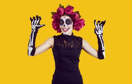 Portrait of a woman with sugar skull makeup, skeleton gloves and flowers on head scaring with hands and smiling isolated on yellow background. Halloween and Day of the Dead concept.の写真素材