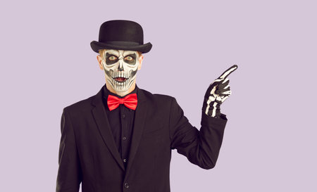 Portrait of man with skull makeup pointing with his finger to side. Young man wearing black suit, bowler hat and skeleton gloves pointing away with index finger at copy spaceの写真素材