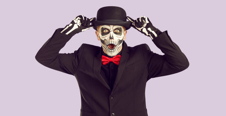 Shocked zomby man holding hat with two hands on lavander background. Black funeral suit with red bow tie and hat - halloween party costume. Skeleton face and body art for day of the dead.の写真素材