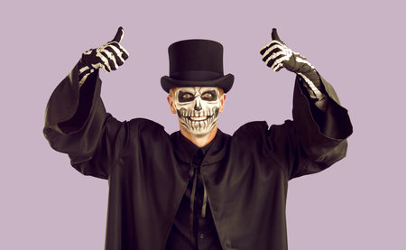 Joyful man in spooky Halloween outfit raise thumbs up recommending festival service. Smiling man with skull make-up wearing cloak and hat recommends something on lilac background. Halloween banner.の写真素材