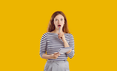 Portrait of red-haired, surprised, shocked, beautiful girl in striped black and white jumper and gray trousers, holding notebook and pen near her open mouth. Studio photo on yellow background.の写真素材