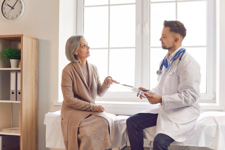 Doctor consulting female senior patient talking, sharing symptoms, complaining, anxious, troubled about health. Old woman visiting clinic for medical care, discussing treatment plan, asking for helpの写真素材