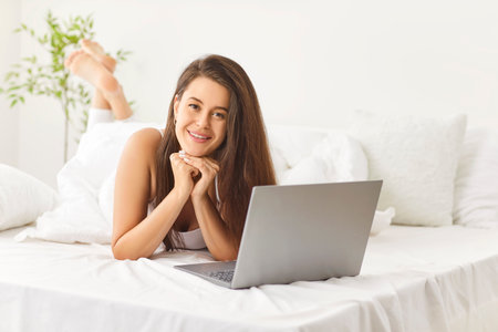 Happy relaxed young woman lying in cozy bed under white blanket with laptop. Smiling caucasian brunette girl looking at camera, enjoying lazy leisure time in bright sunlit bedroom during weekend.の写真素材
