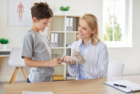 Young smiling woman stomatologist teaching cleaning teeth a little child boy patient for oral health in hospital. Professional dentist teaching how to use toothbrush in dental office.の写真素材