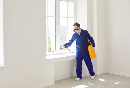 Man exterminator fighting against insects in house spraying poison from sprayer bottle on window. Worker in protective face mask disinfecting apartments or doing pest control with disinfectant sprayの写真素材