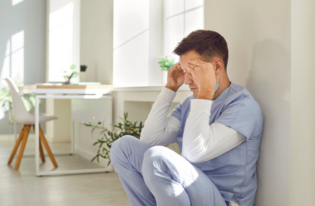 Stressed man nurse or doctor in blue scrubs feeling tired, exhausted and completely burnt out after long difficult work shift under constant pressure sitting by wall on floor holding hands on foreheadの写真素材