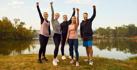 Happy sporty people in sportswear standing with hands up in fist like winners on lake shore after successful sport exercises in park. Outdoors training and fitness in nature concept. Banner.の写真素材