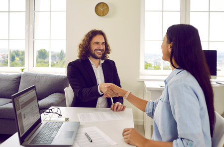 Young happy smiling business man sitting in office at the desk on workplace shaking hands with woman reaching agreement and making a good deal or signing a contract. Business people at work.の写真素材