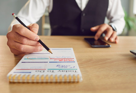 Cropped photo of business man writing tasks and to do list in notepad sitting at the desk with smartphone in hand at home or at office planning daily appointment in plan schedule. Planner concept.の写真素材