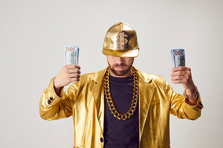 Portrait of funny successful confident young man in golden suit and cap holding money dollars cash in hands isolated on white studio background. Business success and investment concept.の写真素材