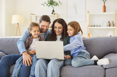 Smiling parents sitting on sofa with two children, hugging, using laptop together. Happy cheerful family watching something fun on screen, enjoying quality time and having fun in living room at home.の写真素材