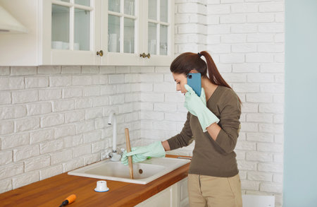 Focused young woman wearing rubber gloves using cup plunger to unclog sink in kitchen while talking on phone. Busy female homeowner calling emergency or plumbing service while solving plumbing issue.の写真素材