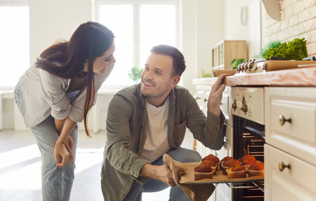 Happy man helping taking tray with yummy self baked tasty muffins out of oven to enjoy cooking in modern kitchen, homemade family pastries, culinary hobby, cupcakes dessert, tasty hot fresh bakeryの写真素材