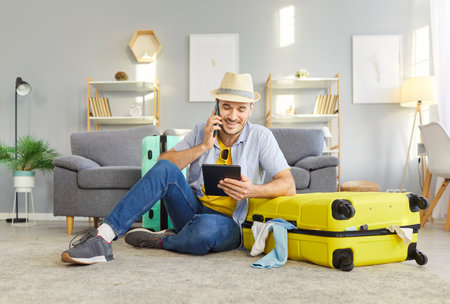 Cheerful male tourist focused at tablet screen for booking hotel room, has phone conversation with friend and planning vacation, posing near packed suitcase in living room. Tourism conceptの写真素材