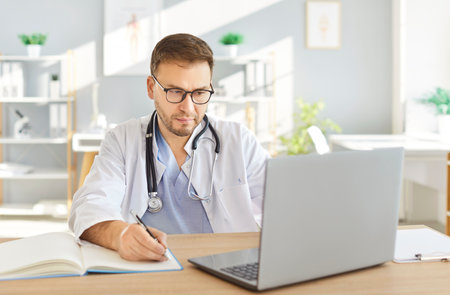 Focused male doctor or nurse working with a laptop and documents in a clinic office. Conducting research, reviewing prescriptions, and handling healthcare tasks with precision and dedication.の写真素材