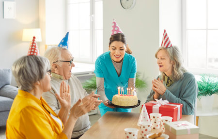 Birthday in nursery house. Nurse and three senior people celebrating anniversary of friend, giving tasty birthday cake with burning candles, enjoying party together, have festive moodの写真素材