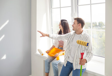 Happy young family couple sitting on window in their new home, holding paint rollers and discussing renovation plan. Cheerful smiling husband and wife preparing to do repair works and painting walls.の写真素材