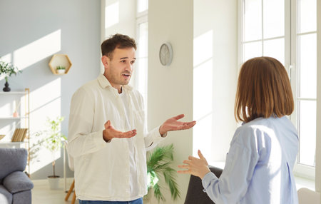 Frustrated young family couple standing by window and arguing with each other. Husband and wife expressing anger and disappointment, struggling with relationship issues and lack of understanding.の写真素材